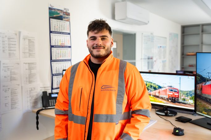 Junger Mann in orangefarbener Arbeitsjacke, lächelt vor einem Büro mit Monitoren.